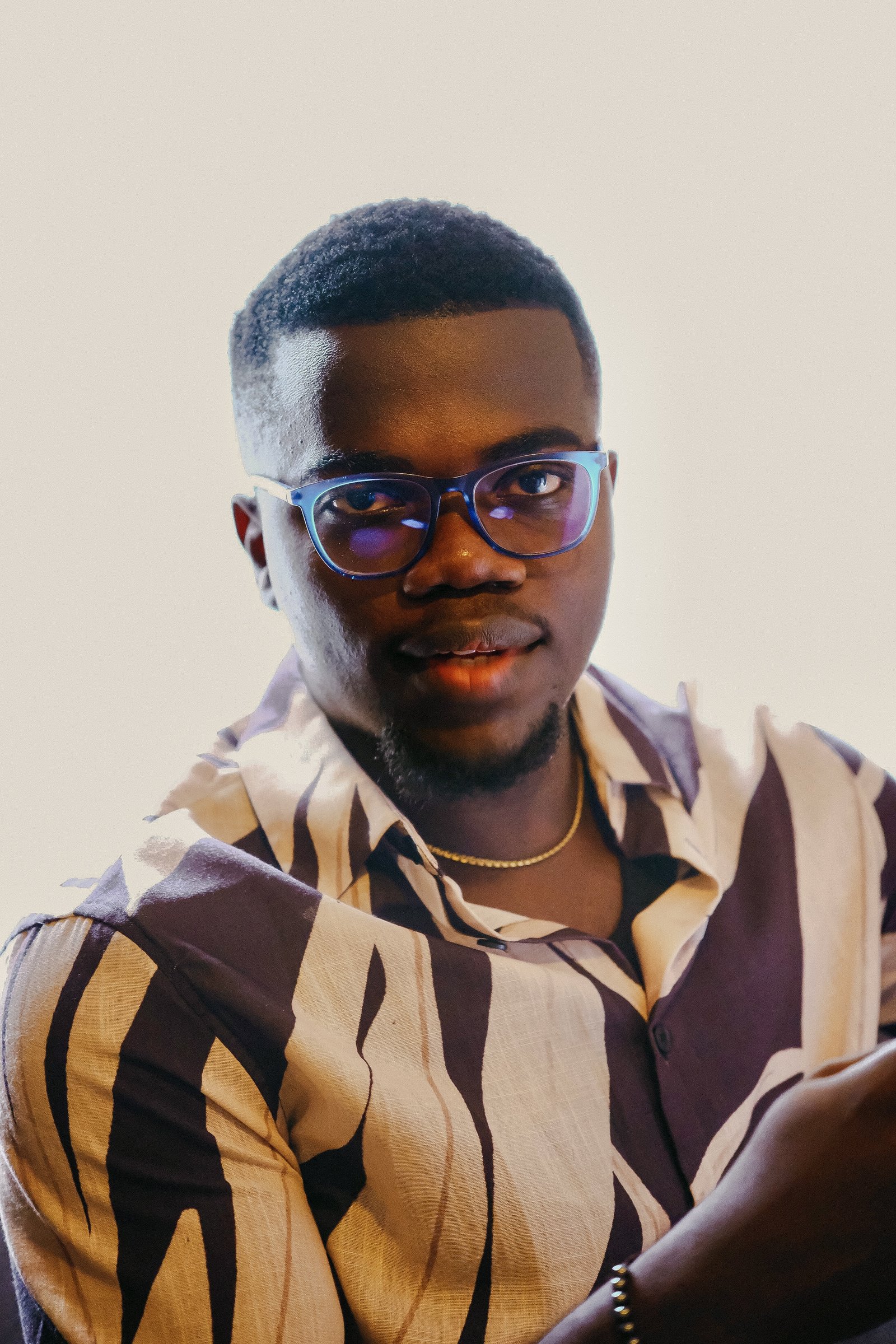 Close editorial portrait of Nnaemeka Nnam wearing blue glasses against a cream background, making direct eye contact