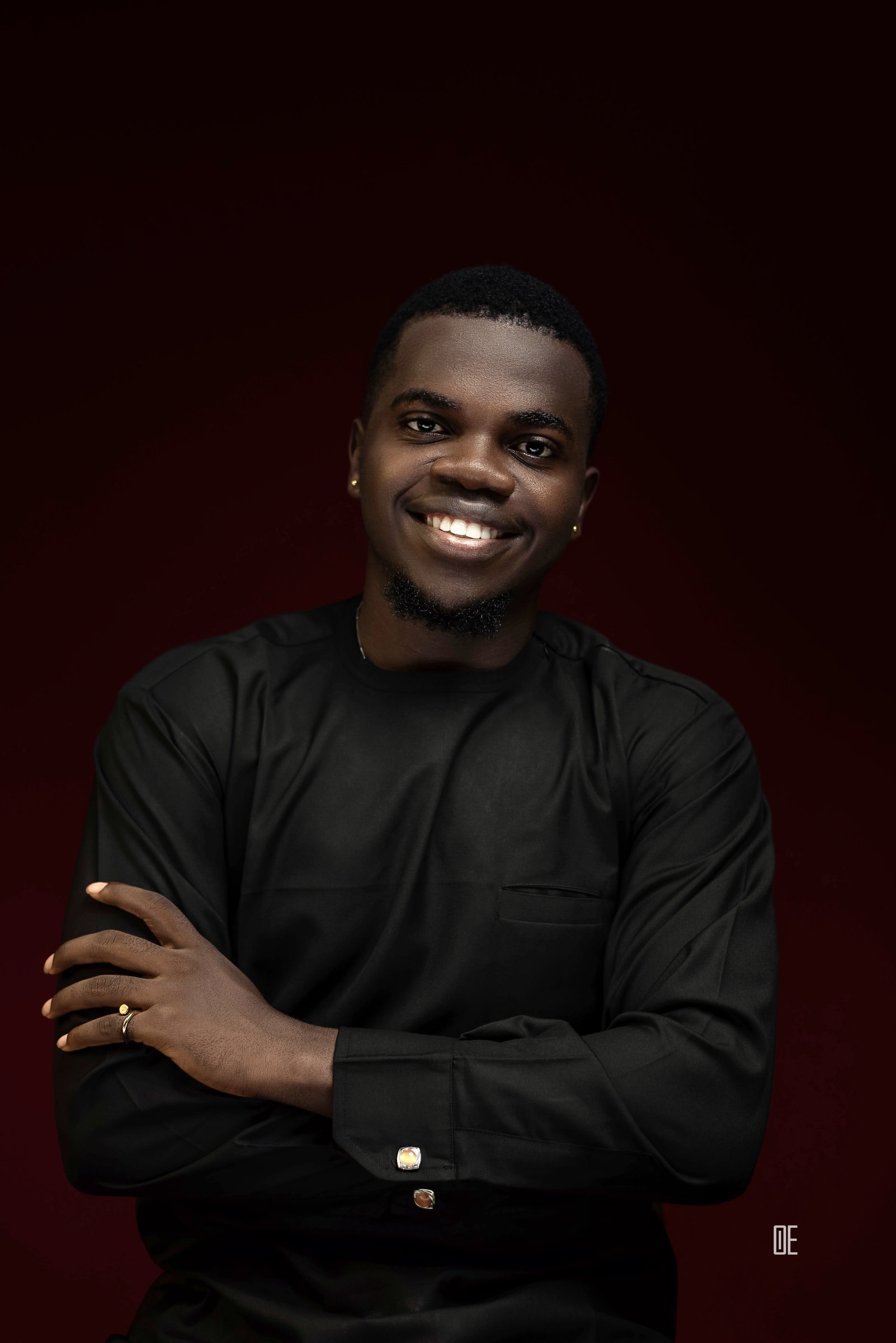 Nnaemeka Nnam in a dark studio portrait, arms crossed, wearing black against a deep maroon background, smiling with confidence