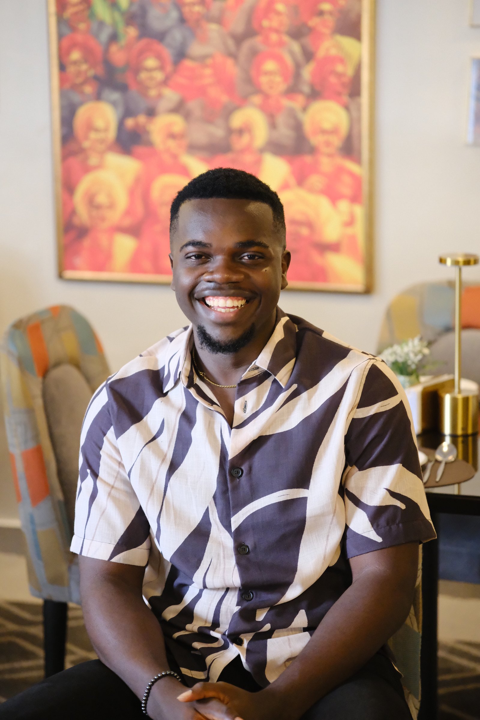 Nnaemeka Nnam seated and smiling broadly in a patterned shirt, with a vivid Nigerian painting on the wall behind him