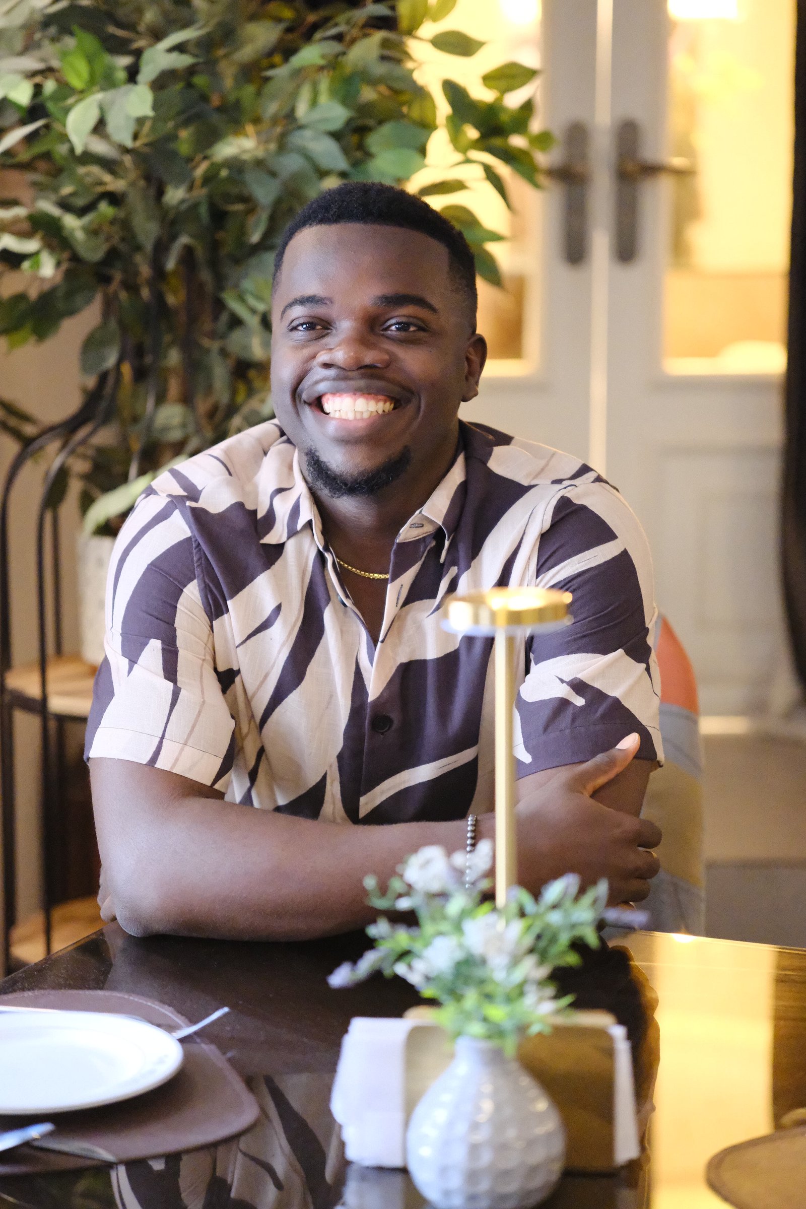 Nnaemeka Nnam with arms folded at a table, smiling warmly, surrounded by greenery and warm light