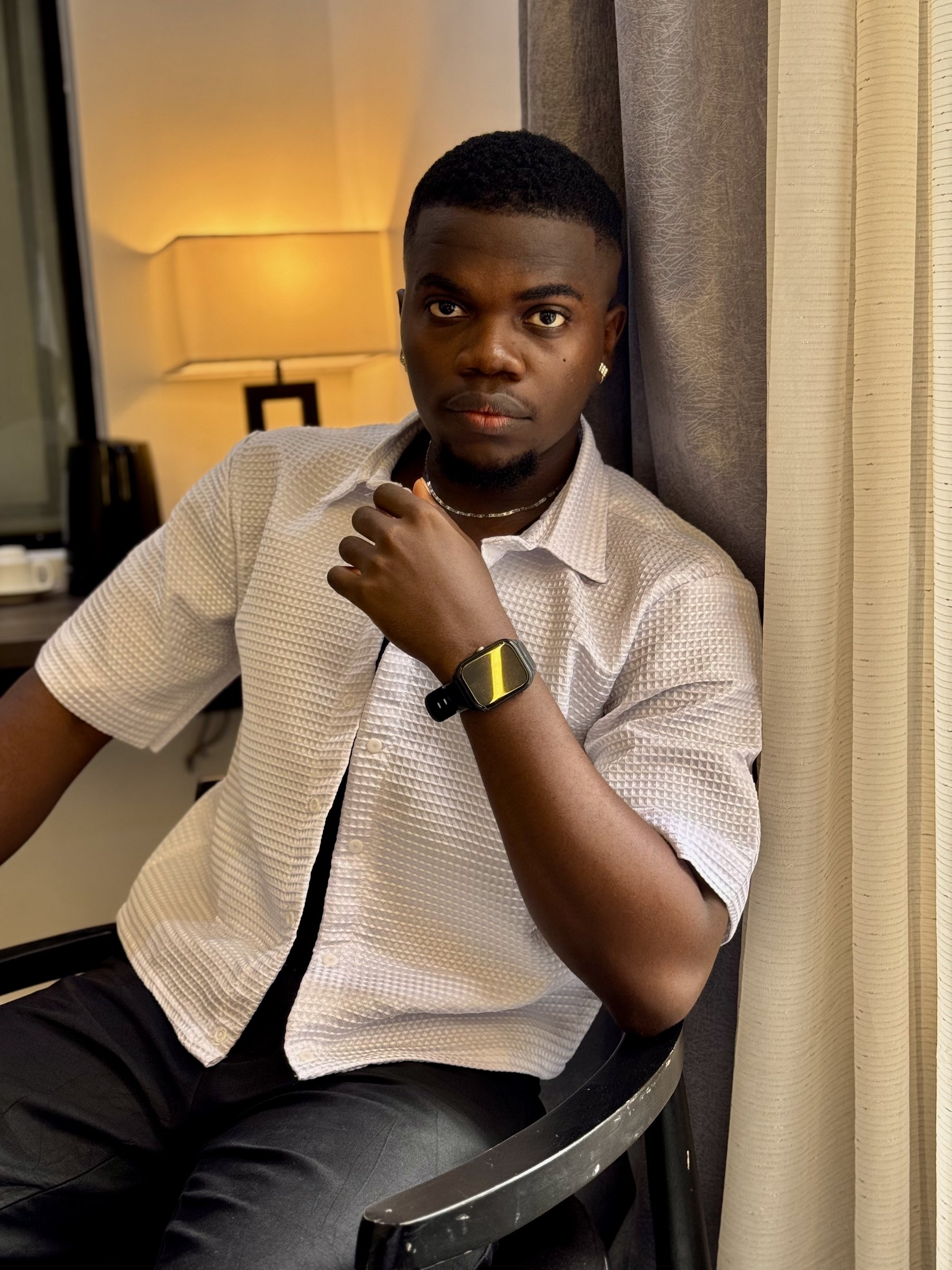 Nnaemeka Nnam seated in a chair in a white textured shirt, looking directly at the camera with a contemplative gaze in moody hotel lighting